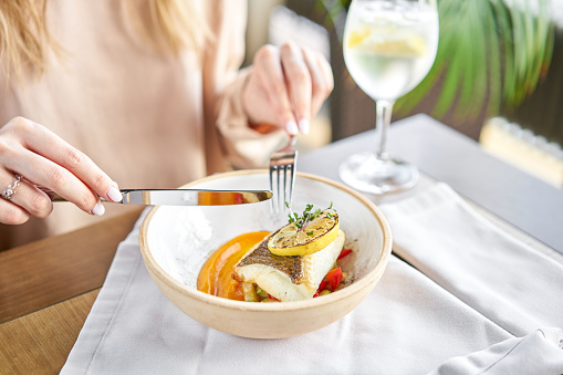 Roasted Halibut with vegetables, paprika pepper and pumpkin cream. Lunch in a restaurant, a woman eats delicious and healthy food. Dish decorated with a slice of lemon. Restaurant menu.