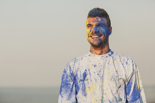 Waist up of happy young man smiling widely while having fun in a colorful festive celebration with blue and yellow powders at the beach.