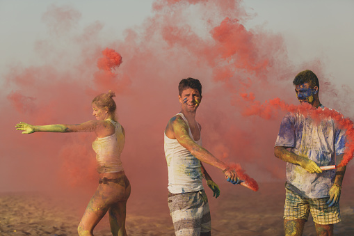 Three quarter length of three lively friends during a summer vacation at the beach playing with powder colors and red smoke flares.