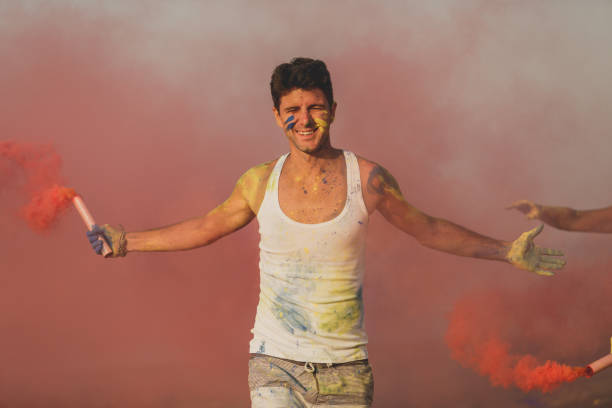 Front view of young man playing with a red smoke flare during a colorful festive celebration at the beach during summer vacations. He opens his arms wide open to enjoy the party.