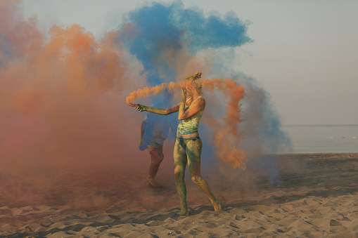 Full length of young woman moving an orange smoke flare during a festive summer celebration at the beach with colorful smoke and powder.