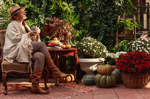Side view of a middle age woman wearing comfortable and warm clothes for autumn, sitting in the outdoors environment of the backyard and surrounded by colorful plants and flowers.