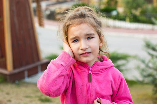 A little girl in a pink hoodie with a sad and tearful face is holding her ear. Ear pain, otitis media, swelling of the cheek, gums, toothache, children's surgery, otolaryngology. Children's medicine