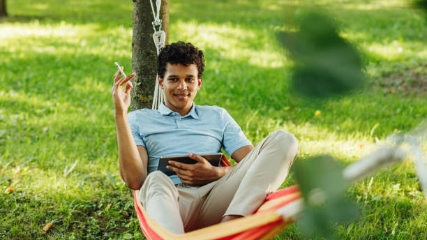 Male student lying on a hammock and holding digital tablet while looking away and thinking Male student lying on a hammock and holding digital tablet while looking away and thinking hammock men lying down digital tablet stock pictures, royalty-free photos & images