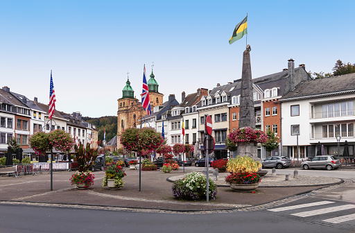 City center with cathedral, obelisk, traditional buildings and flags at the central square of Malmedy in the province of Liege, Wallonia, Ardennes, Belgium.  Cityscape with bright blue sky in autumn season.