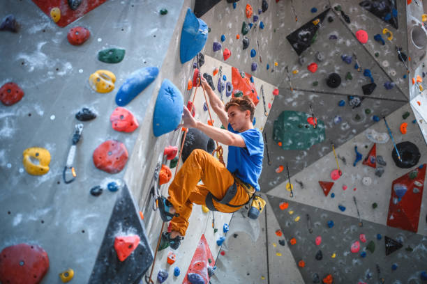 Personal perspective of sportsman in early 20s concentrating on hold route as he scales wall in modern sport climbing center.