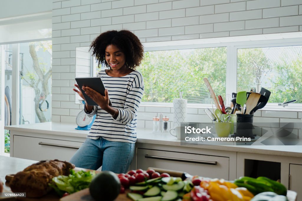African American smiling female researching recipes on digital tablet leaning against counter in clean kitchen Happy African American smiling female researching healthy lunch recipes on digital tablet leaning against counter in modern kitchen. High quality photo Women Stock Photo