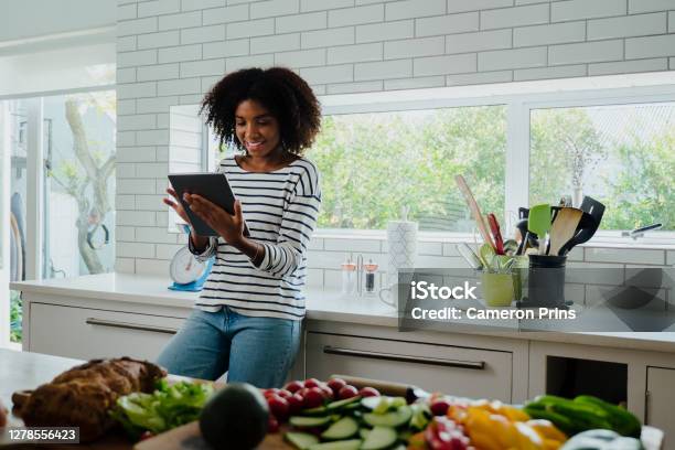 African American Smiling Female Researching Recipes On Digital Tablet Leaning Against Counter In Clean Kitchen Stock Photo - Download Image Now