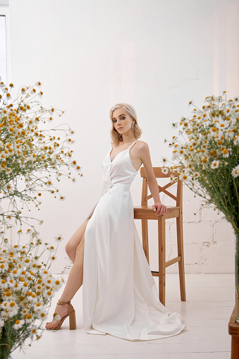 A blonde woman in a beautiful white dress is standing at home among bouquets of wild flowers. Romantic girl with beautiful natural makeup