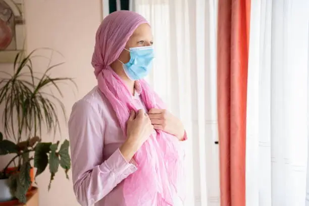 Woman with cancer, pink headscarf and mask looking out the window