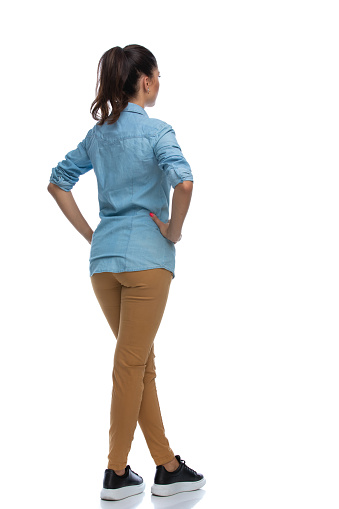 Side rear view of smart casual woman holding hands on hips and standing with legs crossed on white studio background