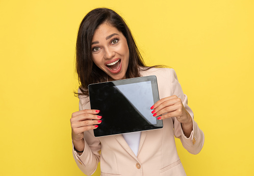 excited young businesswoman in pink suit presenting empty tab screen, opening mouth and smiling in a surprised manner on yellow background