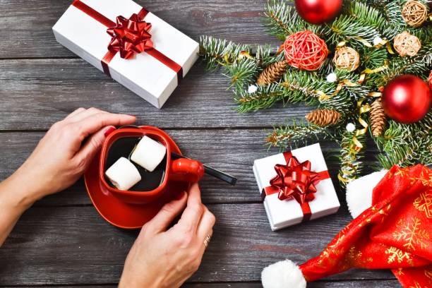 istock Christmas composition. Female hands holdings red cup hot coffee with marshmallow, gift boxes, Santa Claus hat on a wooden background. Christmas concept. Merry Christmas. 1278584592