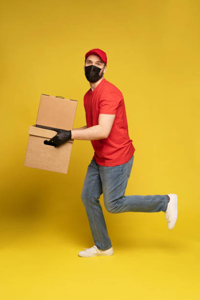 istock Delivery man in protective mask and gloves with box in studio isolated on yellow background. 1278592674