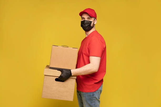 istock Delivery man in protective mask and gloves with box in studio isolated on yellow background. 1278592679