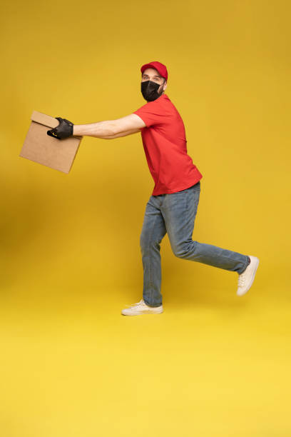istock Delivery man in protective mask and gloves with box in studio isolated on yellow background. 1278592684