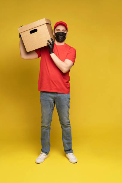 istock Delivery man in protective mask and gloves with box in studio isolated on yellow background. 1278592685
