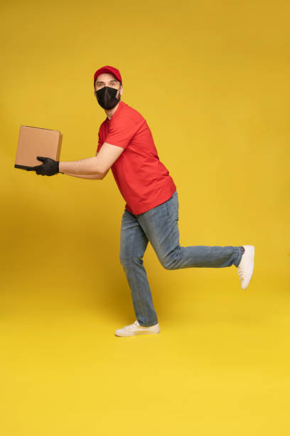 istock Delivery man in protective mask and gloves with box in studio isolated on yellow background. 1278592686