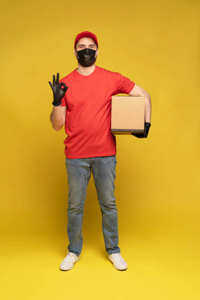 istock Delivery man in protective mask and gloves with box in studio isolated on yellow background. 1278592687