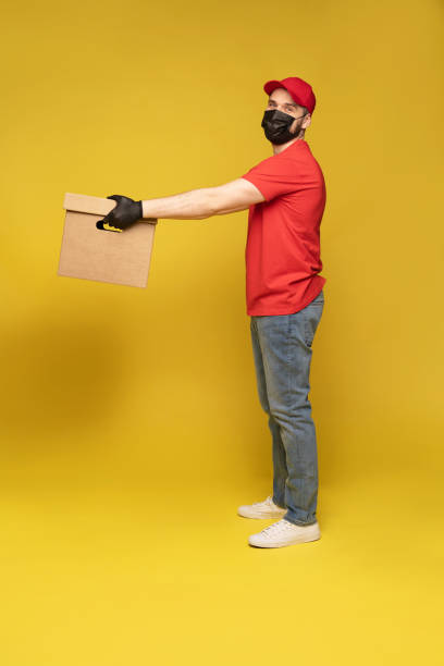 istock Delivery man in protective mask and gloves with box in studio isolated on yellow background. 1278592692