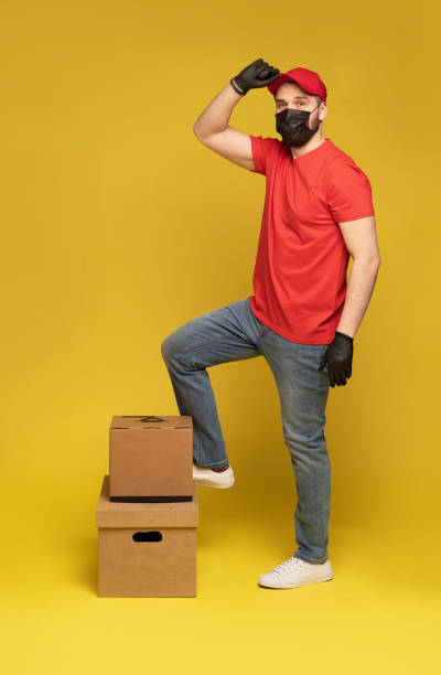 istock Delivery man in protective mask and gloves with box in studio isolated on yellow background. 1278592696