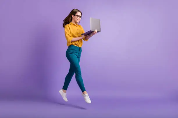 Full length profile photo of lady jump, hold netbook look screen wear yellow shirt blue pants sneakers isolated violet color background