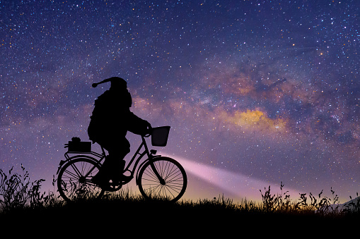 Silhouette of Santa Claus riding on his bicycle to carry a gift under the Milky Way background.