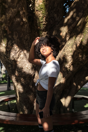 A beautiful African American woman with afro hair looks at the sun under the shade of a lush tree in an outdoor park and holds her cell phone in her hand