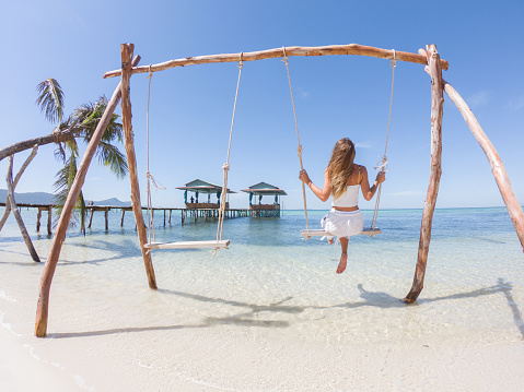 Rear view of woman swinging on white sand beach relaxing and sunbathing by the sea lagoon