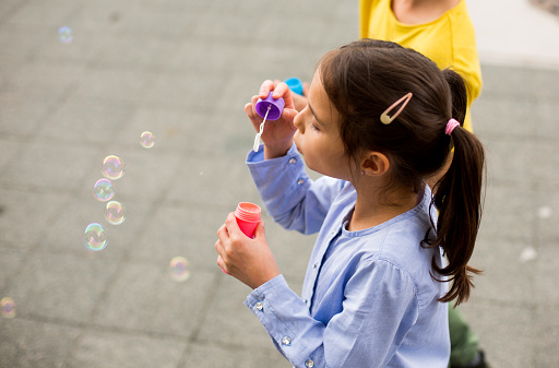 A girl is blowing soap bubbles with her friend. The day is perfect because the bubbles can fly so high.