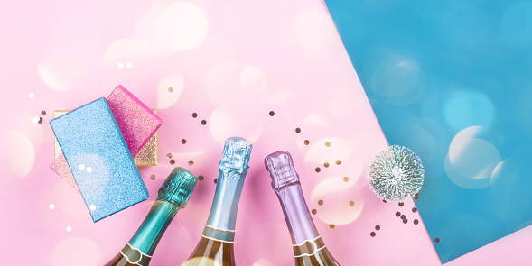 Champagne bottle, gifts and confetti on pink and blue background with sparkling confetti. Celebrating new year, christmas festive flat lay banner
