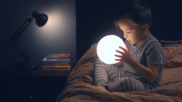 A child is sitting on the bed and holding a glowing moon in his hands.
