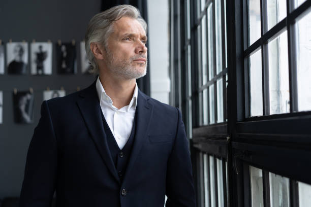 concepto de éxito. elegante hombre barbudo maduro en traje elegante de pie cerca de la ventana. - macho beautiful standing beauty fotografías e imágenes de stock