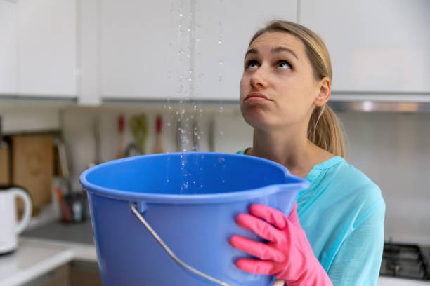 home flooded by upstairs or roof damage - woman holding bucket while water leaking from ceiling in kitchen home flooded by upstairs or roof damage - woman holding bucket while water leaking from ceiling in kitchen property damage stock pictures, royalty-free photos & images