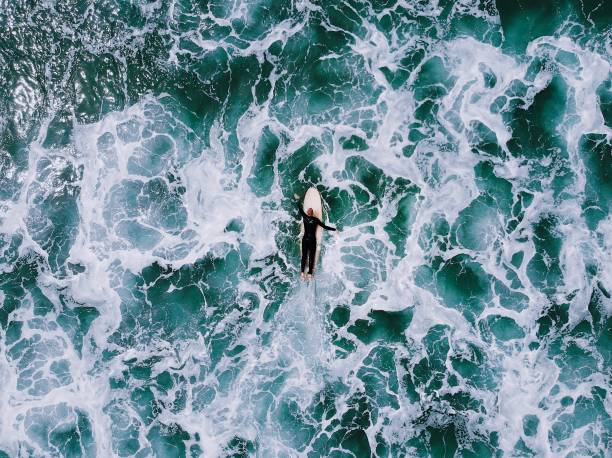 A lone man in a wetsuit paddles out on a handmade wooden surfboard through turbulent white water in blue the ocean Aerial view of rough water where a surfer paddles out through the turbulence surfboard fin stock pictures, royalty-free photos & images