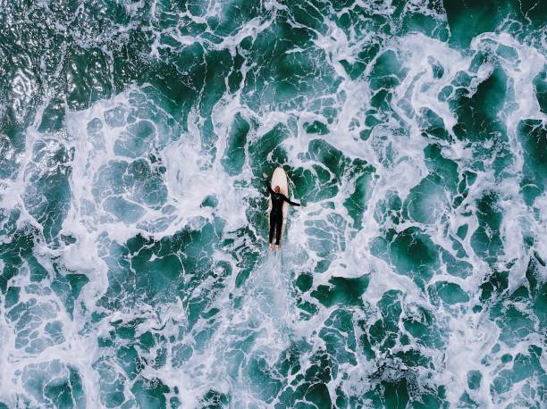 одинокий человек в гидрокостюме весла на ручной деревянной доске для серфинга через бурную белую воду в синем океане - sea water surf tide стоковые фото и изображения
