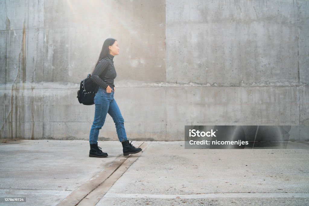 Asian woman walking outdoors against grey wall. Portrait of young beautiful Asian woman walking outdoors against grey wall. 18-19 Years Stock Photo