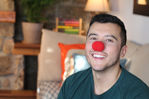 Young ethnic man wearing clown nose at home.