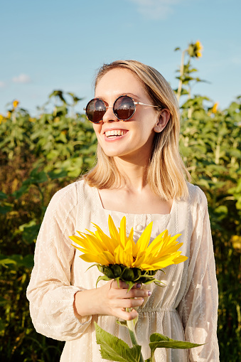 Joyous young female in sunglasses and white dress standing by one of large sunflowers in front of camera in the field against clear sky