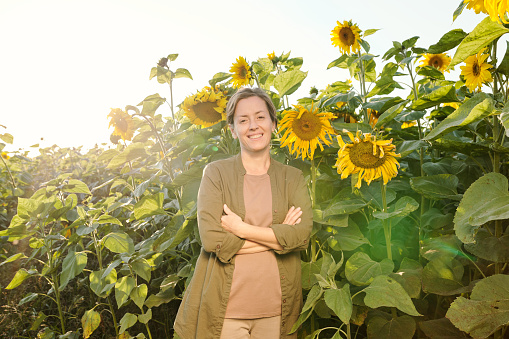 Happy mature woman in casualwear crossing her arms on chest and looking at you while standing in front of camera against sunflowers