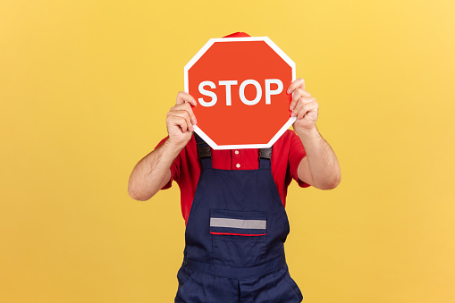 Man in blue overalls and red t-shirt covering face holding stop road sign, warning of danger, restriction and limits. Indoor studio shot isolated on yellow background