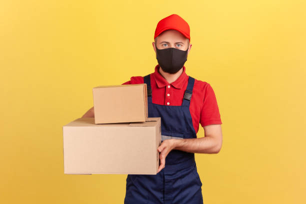 istock Courier in blue overalls red t-shirt and cap and protective mask holding cardboard boxes, express parcels delivery on quarantine 1278637521