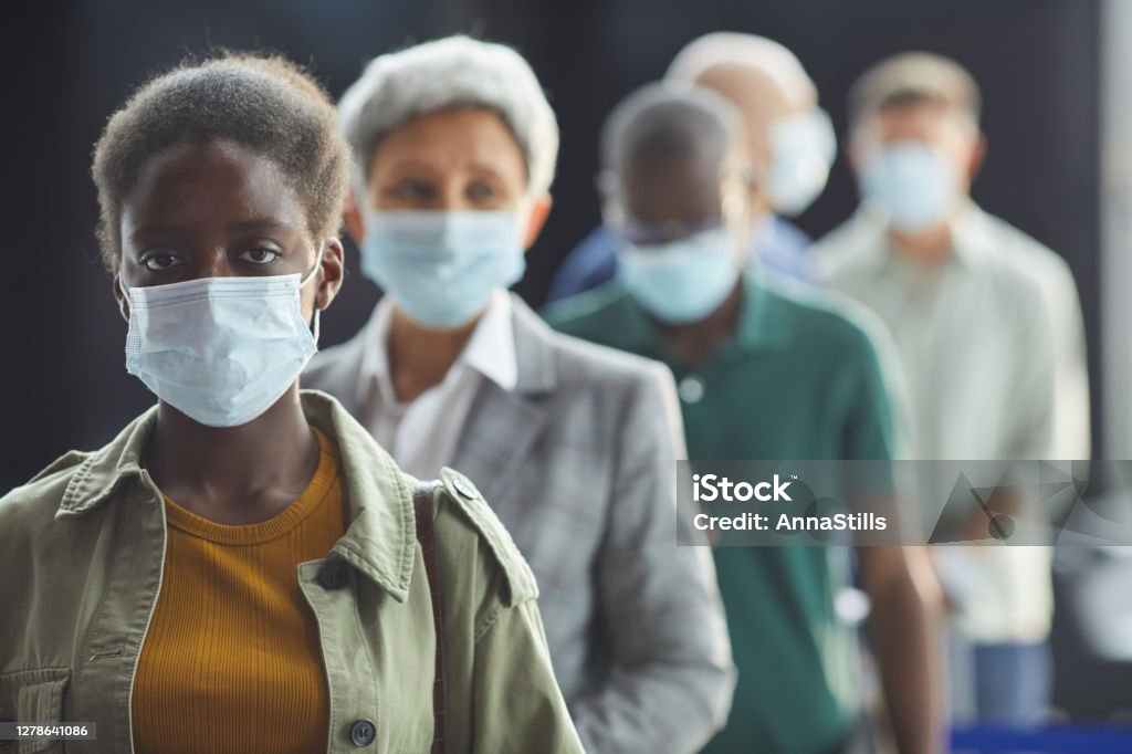 Woman in mask standing in a queue Portrait of young African woman in protective mask looking at camera while standing in a queue Waiting In Line Stock Photo
