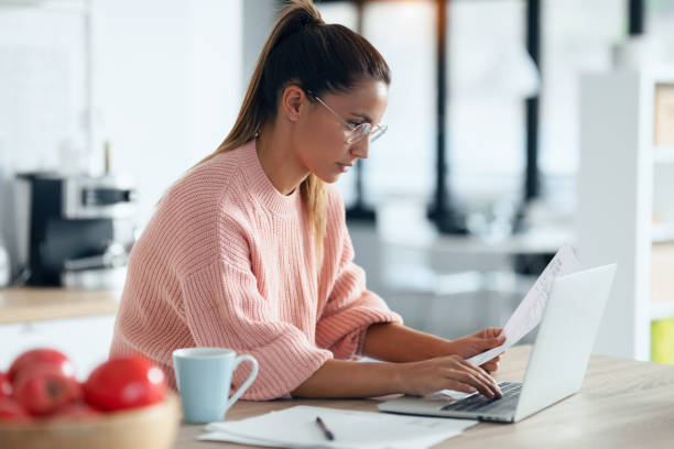istock Concentrated young woman working with computer while consulting some documents in the kitchen at home. 1278651675
