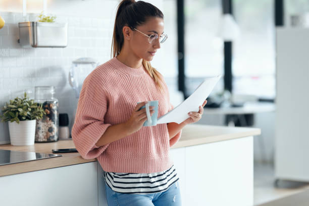 istock Beautiful young business woman drinking a cup of coffee while using her smart phone while holding papers in the kitchen at home. 1278651744