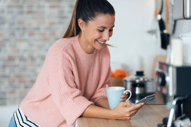 istock Smiling young woman using her mobile phone while drinking a cup of coffee in the kitchen at home. 1278651870