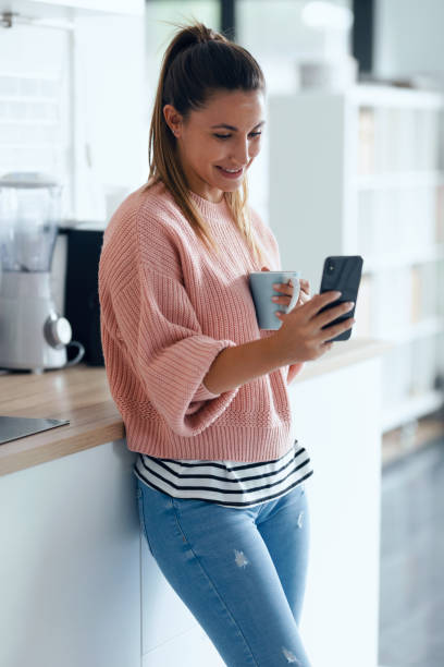 istock Smiling young woman using her mobile phone while drinking a cup of coffee in the kitchen at home. 1278652013