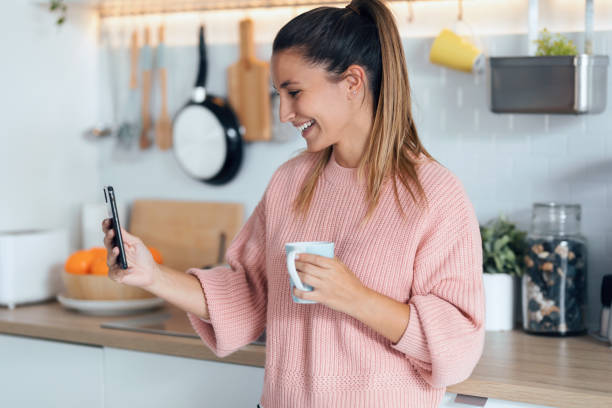 istock Smiling young woman using her mobile phone while drinking a cup of coffee in the kitchen at home. 1278652167