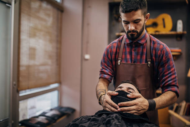 guapo barbero cubriendo la cara del cliente masculino con toalla caliente antes de afeitarse - male towel men human face fotografías e imágenes de stock