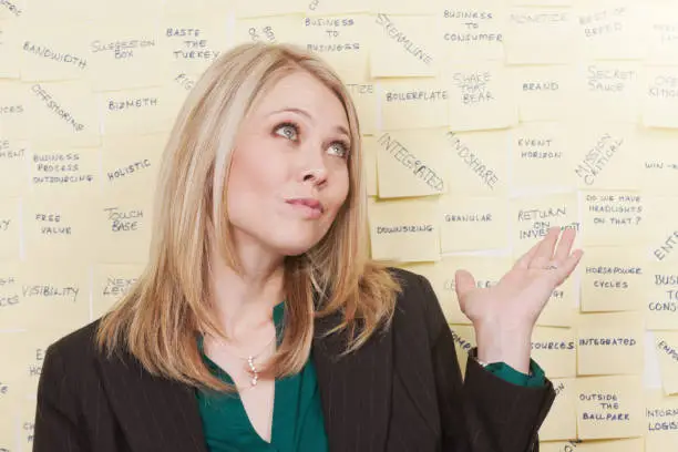 Photo of Young businesswoman shrugs at a wall full of sticky notes inscribed with business buzzwords jargon